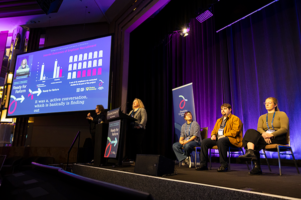 3 people sit on chairs on a stage, 1 person is presenting at a microphone