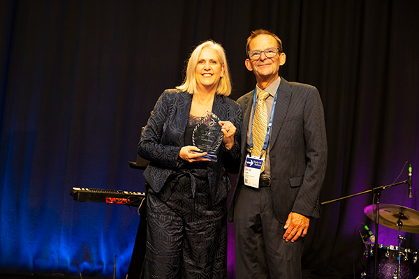 A woman in a black suit with blonde hair holds a trophy next to a man in a suit and tie wearing glasses on stage