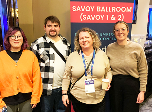 4 people smiling in front of a banner. A woman wears an orange jumper, a man with a beard wears a black and white check shirt, a woman in the front wears a cream top and a women with glasses wears a beige jumper