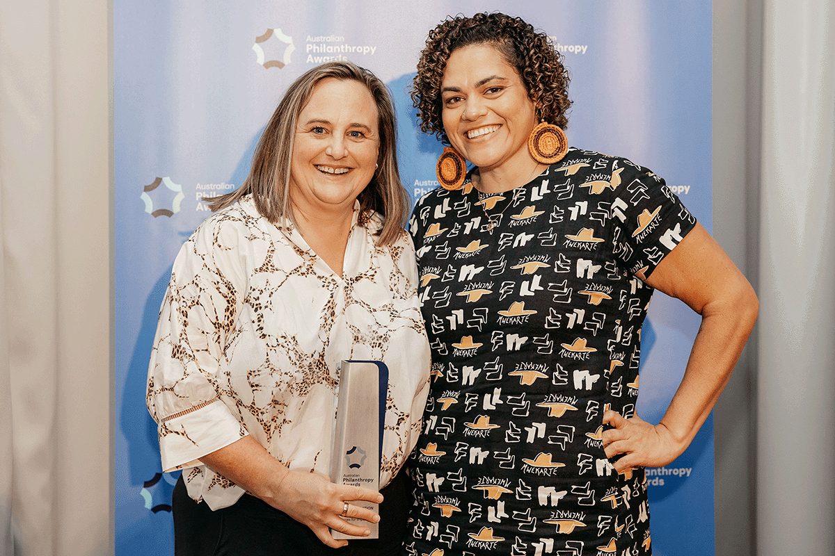 Maree wears a white shirt and is holding a trophy. She is standing next to a woman in a black dress with orange earrings