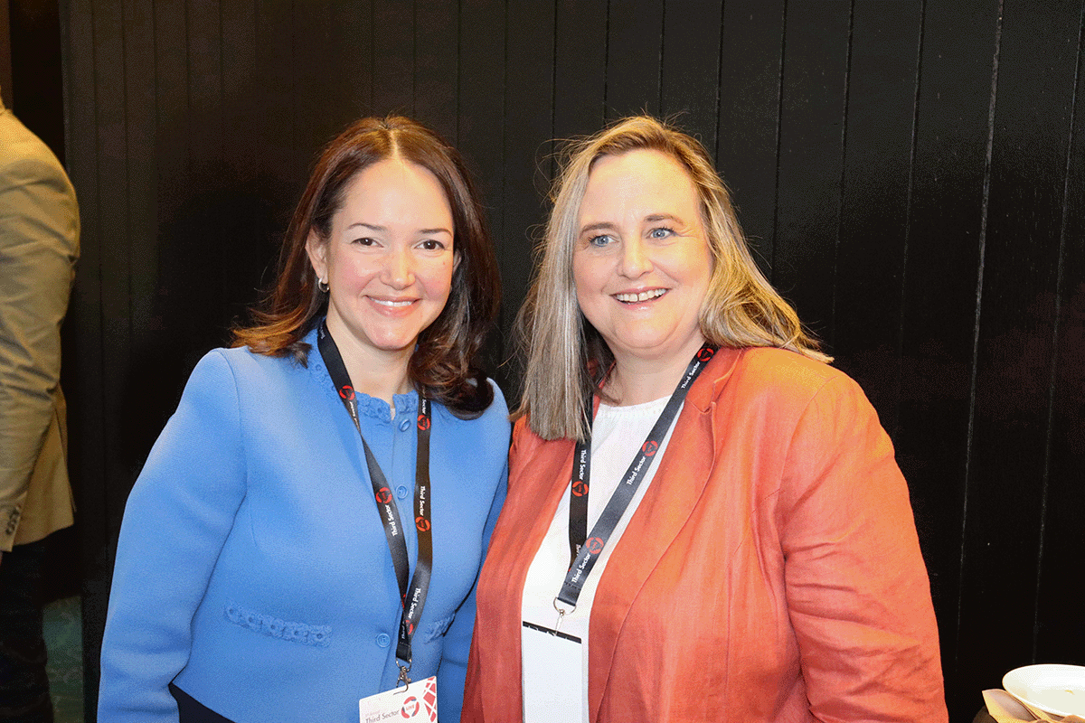 A woman in a blue jacket with brown hair smiles next to a woman with fair hair and an orange jacket