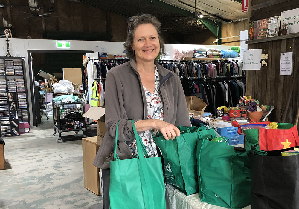 Photo of a woman with a grey jacket, holding a green tote bag. She is smiling at the camera. The background is a store.