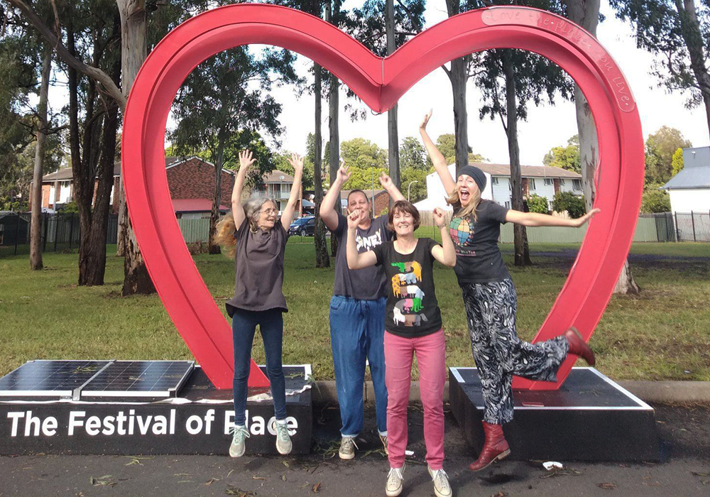 Photo of 3 women and a man standing in front of a sign that says 'The Festival of Place'. They have their arms up and are smiling at the camera.