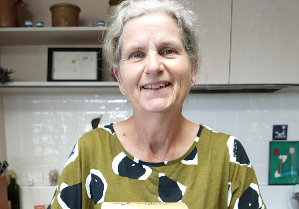 Photo of Sally McCreath. She has grey hair tied back and is wearing a green and black coloured shirt. She is smiling at the camera.