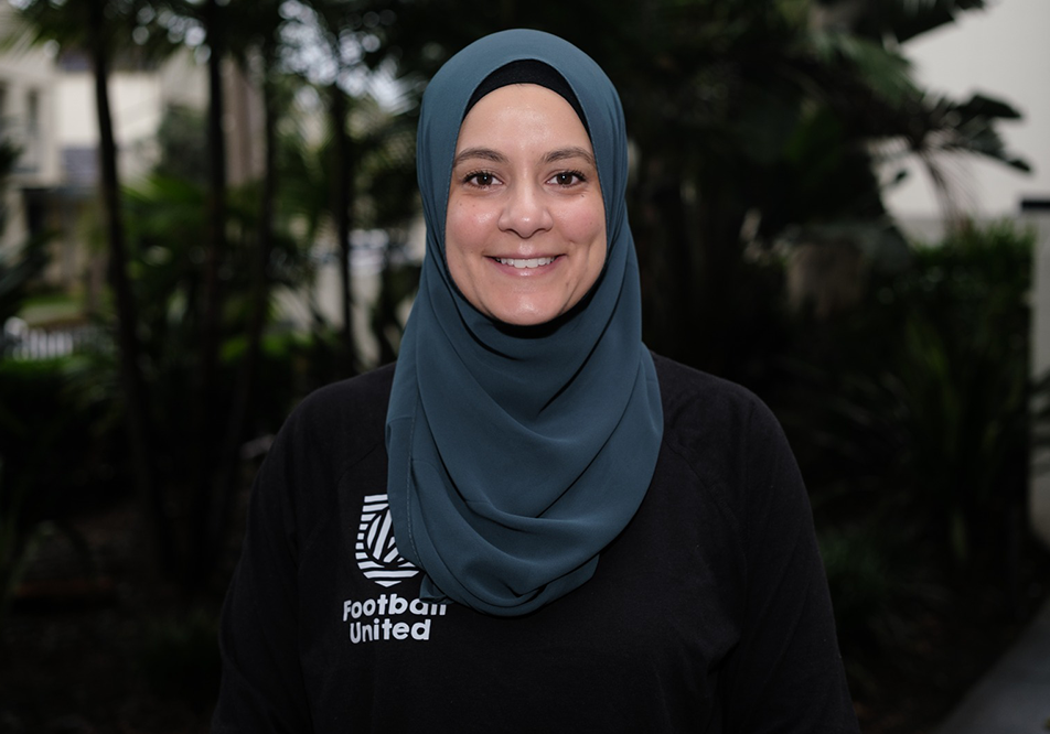 Photo of Assmaah Helal. She is smiling at the camera, wearing a blue headscarf and a black shirt that reads Football United. She is standing in front of a blurred background of green trees.