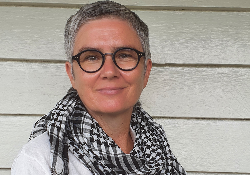 Photo of a person with short hair and glasses, wearing a white top and black checkered scarf. They are smiling at the camera.