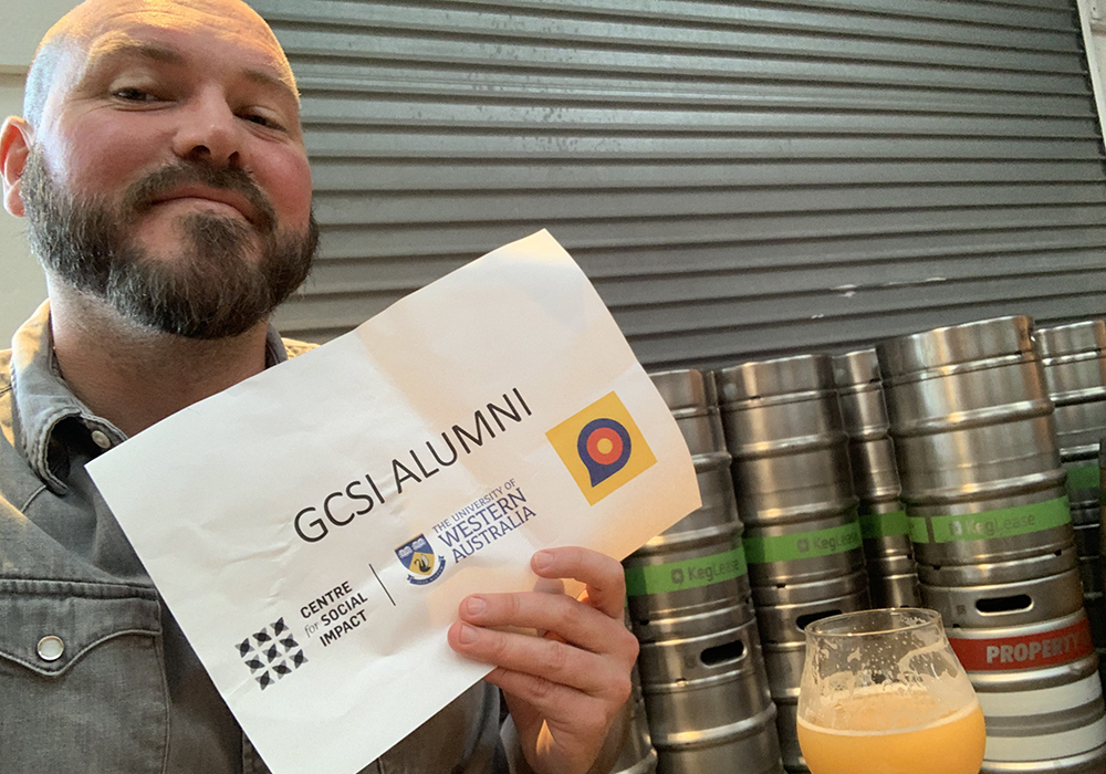 Photo of a man with a beard, wearing a collared shirt. He is holding a sign that says 'GCSI Alumni', and is smiling at the camera.