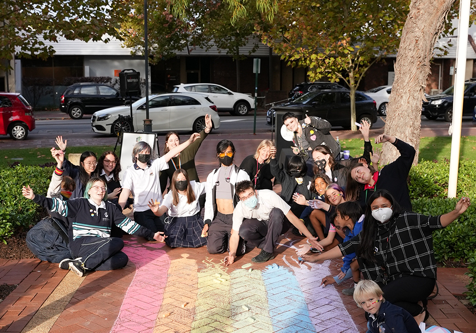 Photo of a group of people sitting in front of a drawing a chalk rainbow. They are smiling at the camera with their arms up in the air.