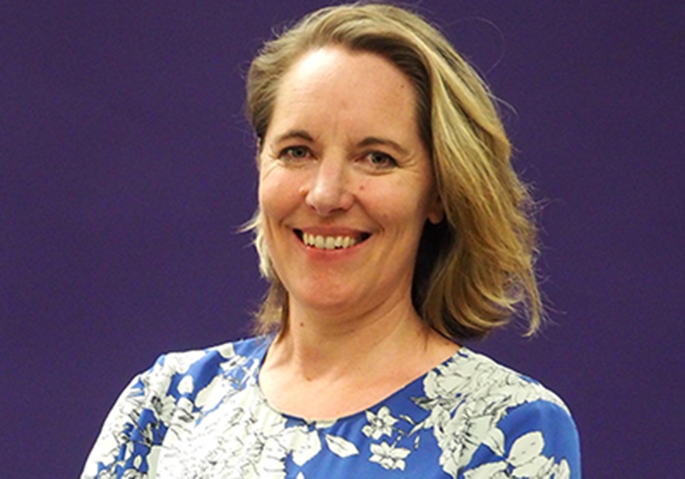 Photo of a woman with light hair, wearing a blue top. She is smiling at the camera.