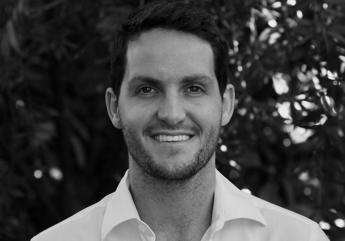 Photo of a man with short dark hair and a beard, wearing a collared shirt. He is smiling at the camera.