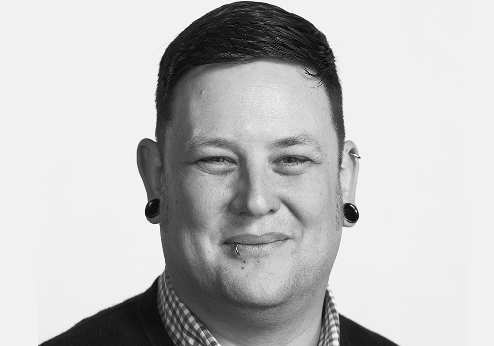Photo of a man with dark hair, wearing black earrings. He is smiling at the camera.