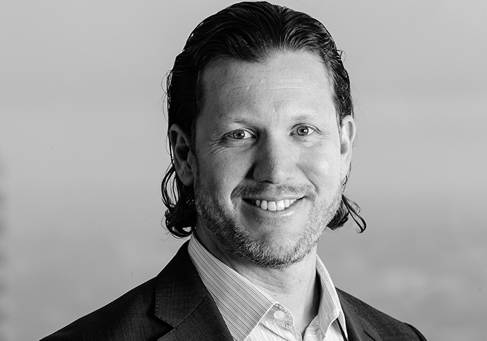 Photo of a man with medium length dark hair and a beard, wearing a collared shirt and jacket. He is smiling at the camera.