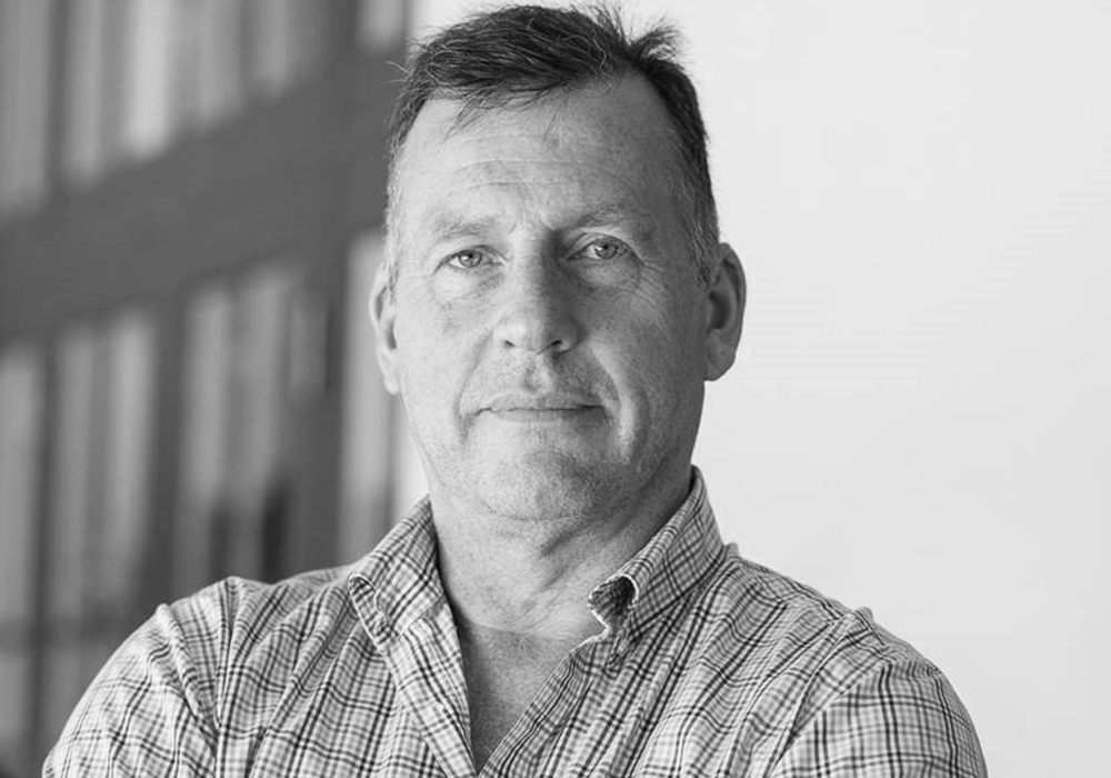 Photo of a man with short dark hair, wearing a checkered collared shirt. He is smiling at the camera.