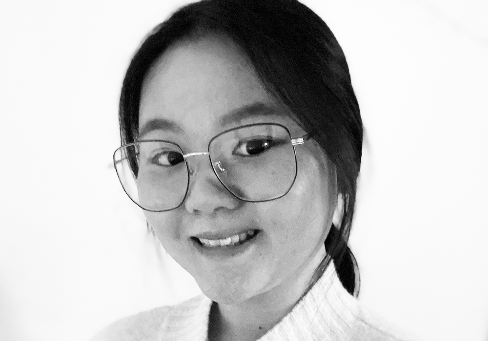 Photo of a woman with dark hair tied up, wearing a light turtleneck and large glasses. She is smiling at the camera.